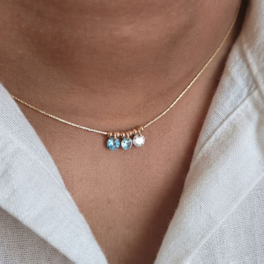 A close-up of a delicate gold chain necklace worn on a person's neck, featuring three small dangling charms: two light blue faceted beads and one white bead.