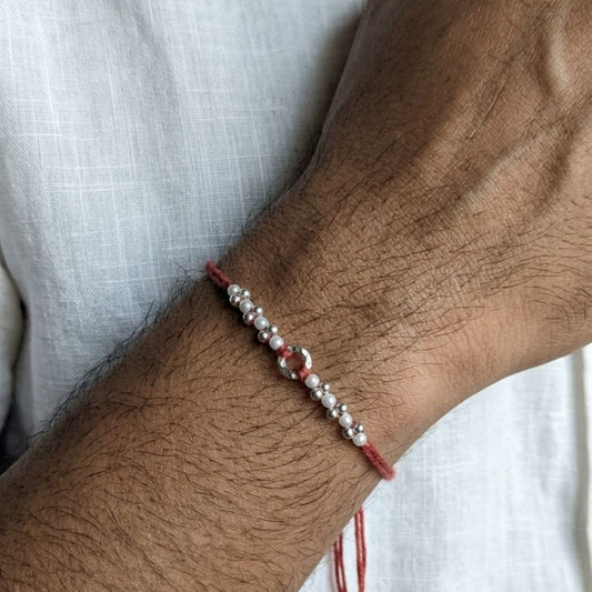 A close-up of a person's wrist wearing a delicate red cord bracelet with silver beads and pearl-like accents. White text labels point to the materials: 'SKIN FRIENDLY COTTON THREAD', 'PEARL', and 'STERLING SILVER'.