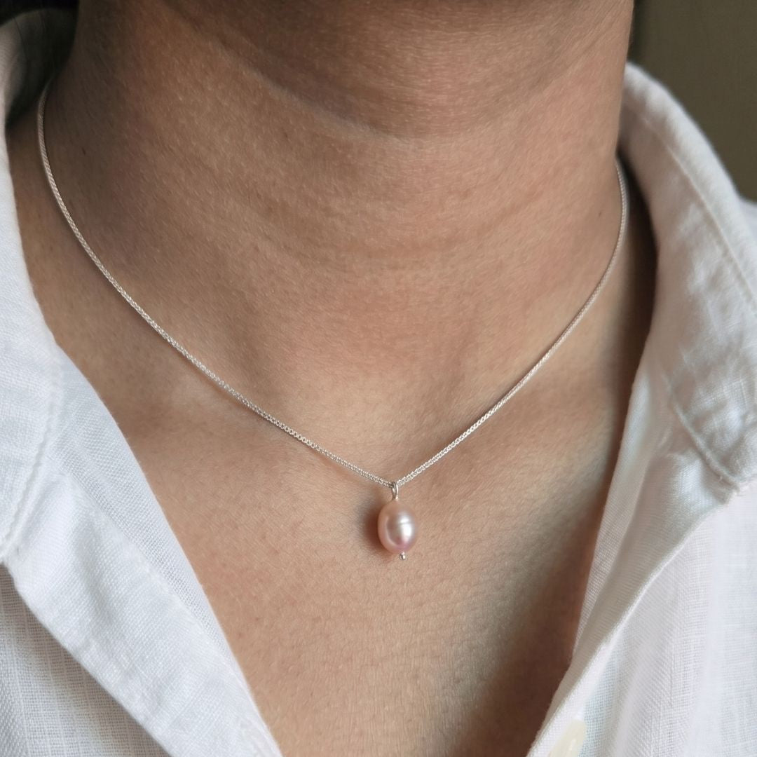 A delicate silver chain necklace featuring a single, teardrop-shaped pink freshwater pearl pendant, shown worn on a person's neck over a white linen shirt.