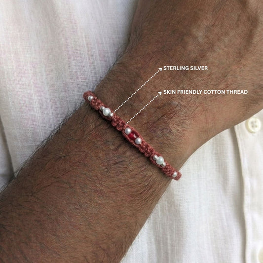 A close-up of a person's wrist wearing a handmade beaded bracelet made of sterling silver beads and red cord, with white text labels pointing out the materials.