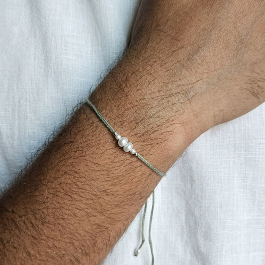 A close-up of a person's forearm wearing a thin gray cord bracelet with three small white pearls at the center, resting against a white textured fabric background.