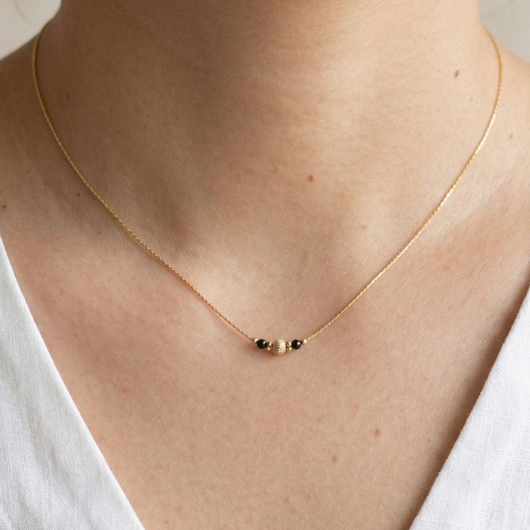 Gold necklace with small black beads on a person wearing a white top, brand 'Minnin' visible.